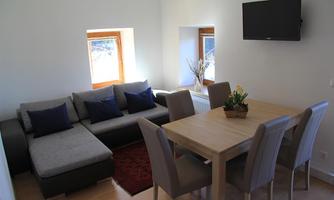 A bright living room with a gray sofa and a wooden dining table. There are large windows that let in daylight.