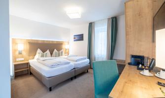 A modern room with two single beds and a desk. The room is bright and friendly, with large windows and tasteful decor.