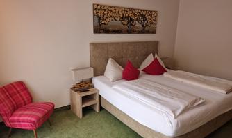 A cozy hotel room with a large bed and red pillows. On it a pink armchair and carpeted floor in a warm tone.