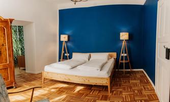 A cozy bedroom with a large bed and two floor lamps. The wall is painted in a bold blue, and the floor is made of wood.
