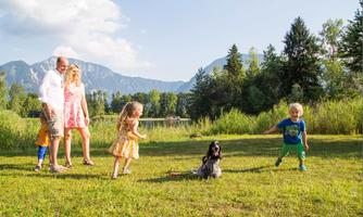 A happy family is playing outdoors with a dog. In the background, there are mountains and trees.
