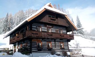 A traditional wooden house with black and white walls in the snow. In the background, there are snow-covered fir trees and a clear sky.