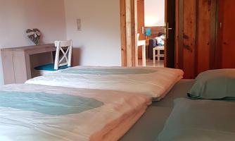 A cozy bedroom with a large double bed and a bright wood design. In the background, a table and an open room are visible.