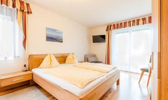 A cozy bedroom with a double bed and light wooden furniture. Large windows let in plenty of light and provide access to a balcony.
