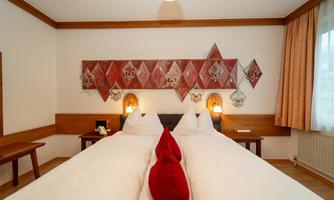 A cozy double room with two beds and red cushions. The walls are adorned with decorative elements and windows that provide natural light.