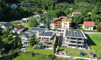 A modern residential complex with several buildings and solar panels on the roofs. Surrounded by green nature and other houses.