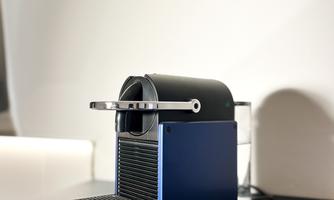 A modern coffee capsule machine in black and silver stands on a table. Next to it, three purple coffee capsules lie on a small plate.