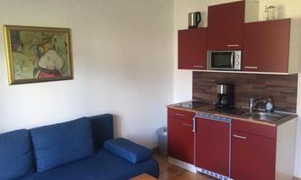 A small kitchen with red cabinets and modern appliances. Next to it is a blue sofa and a wooden table.