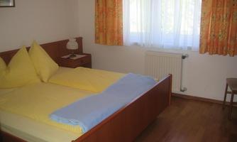 A cozy bedroom with a large double bed and colorful curtains. There is a nightstand, a chair, and plenty of daylight.