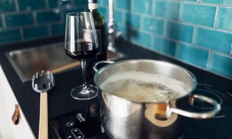 A cooking pot with boiling water is on the stove. Next to the pot stands a glass of red wine and a bottle.