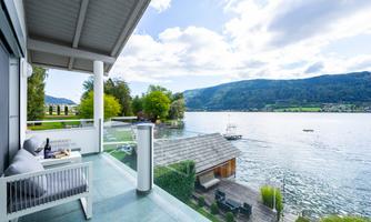 A modern terrace with a view of a tranquil lake and green hills in the background. The clouds are partly cloudy and nature is peaceful.