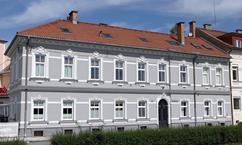 An elegant, historic building with a gray facade and red roof tiles. The facade is adorned with decorative elements and many windows.