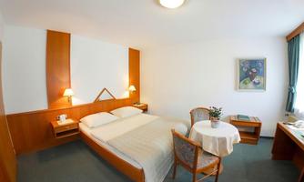 A simple bedroom with a double bed, a table, and chairs. The wall is painted in light colors, and a painting hangs above it.