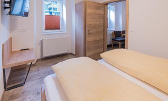 A modern bedroom with two beds and wooden flooring. The room features a television and a window with daylight.