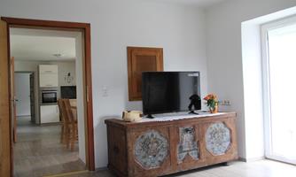A bright living room with an old wooden cabinet and a television. In the background, the kitchen is visible, and the windows allow plenty of daylight in.