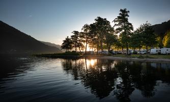 A calm lake with reflections of trees and gentle hills in the background. The sun rises behind the trees, casting a warm light over the scene.