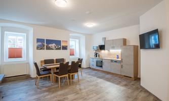 A modern kitchen with a dining area. The walls are light and there are windows with shutters.