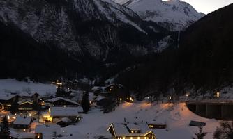 A winter landscape with snow-covered mountains and illuminated houses. The scene is tranquil and inviting at dusk.