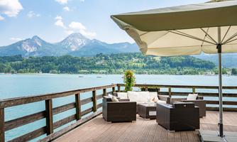 A cozy terrace by the water with modern furniture and a sunshade. In the background, green mountains and a blue sky can be seen.