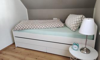 A bedroom with a white bed and a light-colored ceiling. Next to the bed is a bedside table with a lamp and a small clock.