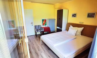 A bright bedroom with a large bed and a cozy armchair. The walls are painted in soft yellow tones and there are decorative pictures.