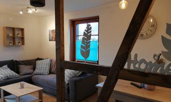 A cozy living space with a gray sofa and a wooden table. There is a window with a view of the street and some decorative elements on the walls.