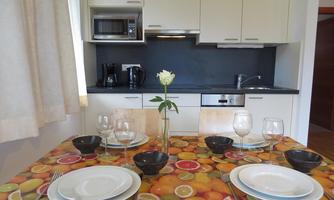 A modern kitchen with a dining table decorated with a colorful fruit tablecloth. The table is set with dishes, glasses, and a white rose in the center.
