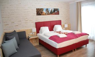 A modern bedroom with a red bed and a gray sofa. The walls are designed in light tones, and a small picture hangs above the bed.