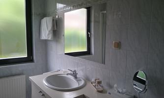 A bright bathroom with a sink and a large mirror. The window has darkened panes and several towels are hanging on the wall.