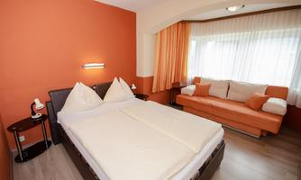 A cozy bedroom with a large bed and an orange sofa. The walls are painted in warm tones and there is plenty of daylight.