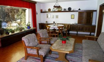 A cozy living room with wooden furniture and a large window. A dining area with a table and chairs is also visible.