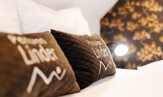 A cozy bed with brown cushions that feature the inscription "Pension Lindner." In the background, there is a stylish wall with a floral pattern.
