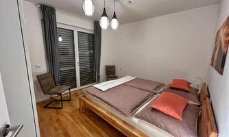 A modern bedroom with a large double bed and cozy pillows. There is a small chair and large windows that let in light.