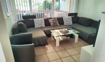 A cozy living area with a large gray corner couch and a glass table. The room is bright and welcoming, with plenty of daylight.