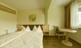 A bright and modern room with two beds, a chair, and a table. Large windows provide pleasant lighting.