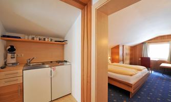 A cozy room with a bed and a small kitchen area. The walls are made of wood, and there is plenty of daylight.