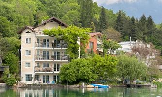 A picturesque building by the water, surrounded by greenery and trees. In the background, mountains and wooded hills are visible.