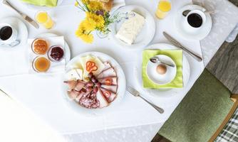An inviting breakfast with cold cuts, cheese, fresh rolls, and jam. The table also features coffee and orange juice, surrounded by colorful flowers.