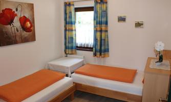 A simple room with two single beds and orange bedspreads.  
Next to a window with colorful curtains, there is a small table and a flower decoration.