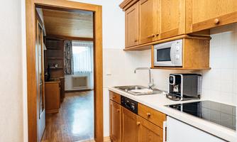 A modern kitchen with wooden furniture and white appliances. In the background, a bright living room with windows is visible.
