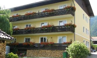 A yellow building with three floors and balconies decorated with blooming plants. In the foreground, piles of wood and green bushes can be seen.