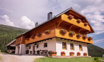 A large wooden house with a balcony decorated with flowers. In the background, green hills and a blue sky can be seen.