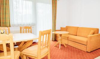 A bright living space with an orange sofa and a table with chairs. The large windows let in plenty of light and the carpet has a classic pattern.