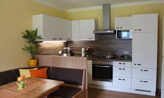 A modern kitchen with a wooden countertop and white cabinets. A dining table with colorful cushions and a plant creates a cozy atmosphere.