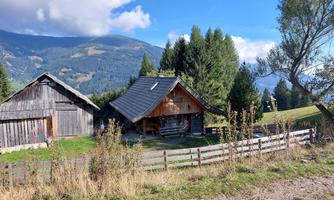 A picturesque alpine hut surrounded by trees and gentle hills. The sky is clear and the landscape is green and inviting.