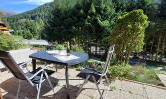 A cozy outdoor area with a table and two chairs. In the background, lush greenery and mountains can be seen under a blue sky.
