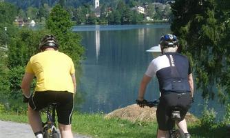 Two cyclists ride along a path overlooking a lake and a picturesque landscape. In the background, a church is visible, surrounded by green hills.