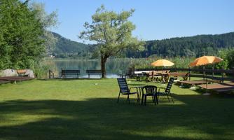 A green meadow by the shore of a calm lake, surrounded by trees. On the grass, there are tables and chairs as well as umbrellas providing shade.