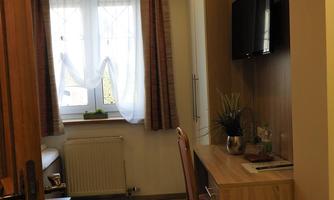 A small room with a table, a chair, and a television on the wall. The window is fitted with white curtains and lets in a lot of light.