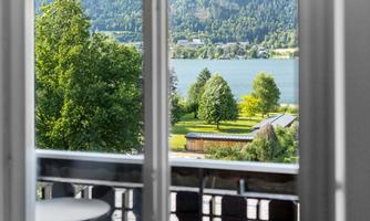 A beautiful view through a window of a clear lake, surrounded by green trees. In the background, gentle hills and buildings can be seen.